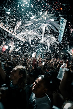 A lively crowd is celebrating under a massive indoor installation with bright, decorative elements hanging from the ceiling. Confetti is falling everywhere, and people are cheering and holding up their phones, capturing the moment.