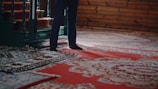 A person stands next to intricately patterned, colorful carpets with a focus on their feet. The carpets feature elaborate designs in shades of red, blue, and white. A wooden staircase with dark green railings is partially visible, along with wooden paneled walls that add a rustic charm to the setting.