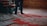 A person stands next to intricately patterned, colorful carpets with a focus on their feet. The carpets feature elaborate designs in shades of red, blue, and white. A wooden staircase with dark green railings is partially visible, along with wooden paneled walls that add a rustic charm to the setting.
