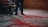 A person stands next to intricately patterned, colorful carpets with a focus on their feet. The carpets feature elaborate designs in shades of red, blue, and white. A wooden staircase with dark green railings is partially visible, along with wooden paneled walls that add a rustic charm to the setting.