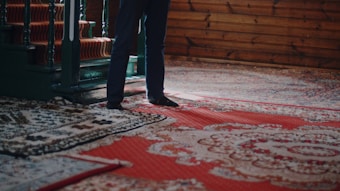 A person stands next to intricately patterned, colorful carpets with a focus on their feet. The carpets feature elaborate designs in shades of red, blue, and white. A wooden staircase with dark green railings is partially visible, along with wooden paneled walls that add a rustic charm to the setting.