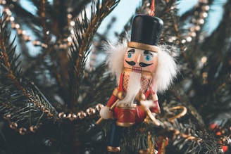 A festive nutcracker ornament is hanging among the branches of a Christmas tree, adorned with beads and other decorative elements. The nutcracker is detailed with a red and gold uniform, white hair, and a black hat.