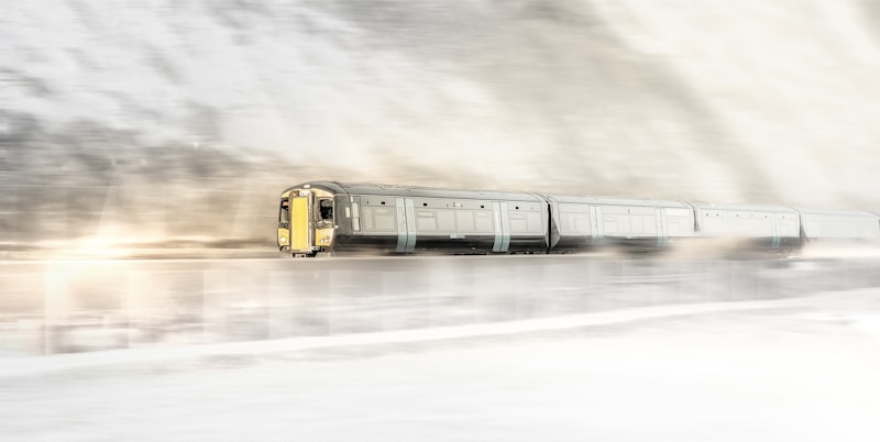 A sleek, modern train travels at high speed through a snowy landscape, creating a sense of motion. The snow flies up around the train, adding to the dynamic feel. The train is silver with yellow accents, and the background is blurred to emphasize movement.