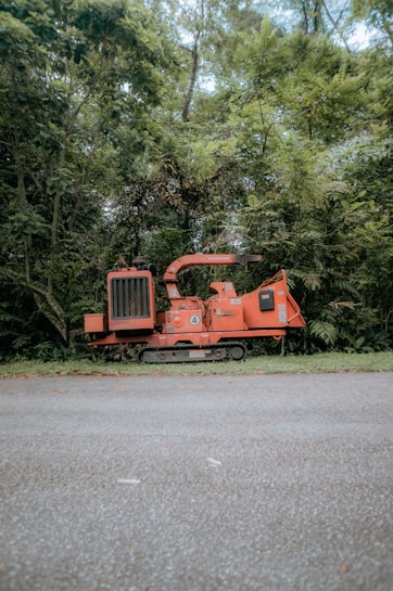 A red piece of industrial machinery, possibly a wood chipper, is situated on a paved pathway surrounded by dense green foliage. Tall trees and lush vegetation create a natural backdrop. The machinery has noticeable metal parts and a chute extending outward.