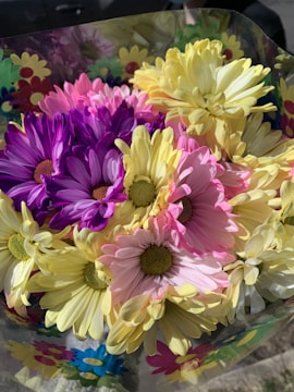 A vibrant bouquet of flowers consisting mainly of daisies in various shades of purple, pink, and yellow. The bouquet is wrapped in a transparent cellophane with colorful flower prints.