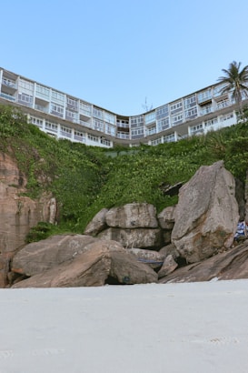 A sandy beach with large rocks and boulders in the foreground. Behind the rocks is a lush green hill covered with vegetation. At the top of the hill, there is a building with numerous windows, likely apartments or offices. A single palm tree is visible to the right.