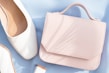 A pastel pink handbag rests on a light blue surface beside a pair of elegant white shoes. The leather handbag has a smooth finish and a subtle embossed logo. The shoes appear to be made of white leather with a faint checkered texture. Soft natural light casts gentle shadows across the items.