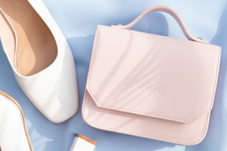 A pastel pink handbag rests on a light blue surface beside a pair of elegant white shoes. The leather handbag has a smooth finish and a subtle embossed logo. The shoes appear to be made of white leather with a faint checkered texture. Soft natural light casts gentle shadows across the items.