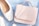 A pastel pink handbag rests on a light blue surface beside a pair of elegant white shoes. The leather handbag has a smooth finish and a subtle embossed logo. The shoes appear to be made of white leather with a faint checkered texture. Soft natural light casts gentle shadows across the items.