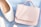 A pastel pink handbag rests on a light blue surface beside a pair of elegant white shoes. The leather handbag has a smooth finish and a subtle embossed logo. The shoes appear to be made of white leather with a faint checkered texture. Soft natural light casts gentle shadows across the items.