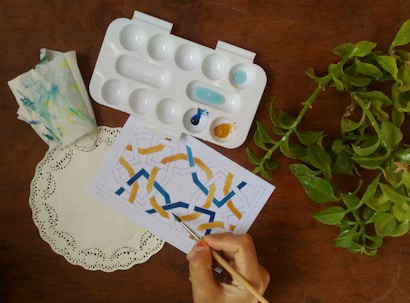 A hand is painting a geometric pattern on a piece of paper using a small brush. An artist’s palette with blue and yellow paint is nearby, along with a paper doily and a piece of cloth with paint stains. Nearby, a leafy green plant adds a touch of nature to the scene on the wooden surface.