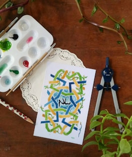 An artistic setup featuring a geometric watercolor painting with shades of blue, green, and yellow, and the word 'Peace' in Arabic. Surrounding the art are painting supplies including a palette with various paints, two brushes, a pencil with ladybug motifs, and a drawing compass. A doily is placed under the painting, and there are green leafy plants nearby.