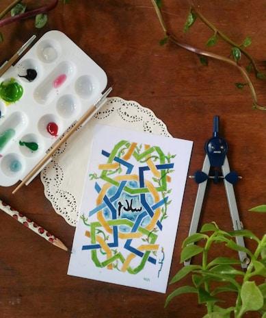 An artistic setup featuring a geometric watercolor painting with shades of blue, green, and yellow, and the word 'Peace' in Arabic. Surrounding the art are painting supplies including a palette with various paints, two brushes, a pencil with ladybug motifs, and a drawing compass. A doily is placed under the painting, and there are green leafy plants nearby.