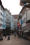 A narrow cobblestone street lined with buildings featuring traditional architecture. Swiss flags are prominently displayed, and several pedestrians, including a person pushing a stroller, are walking down the street. The scene conveys a quaint, European urban atmosphere.