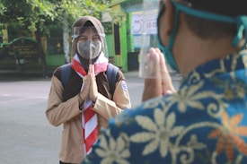 A person wearing a scout uniform is standing with hands in a greeting gesture, wearing a face mask and a transparent face shield. Another person, partially visible, is facing them and also using a similar greeting gesture. The setting appears to be outdoors with green foliage and buildings in the background.