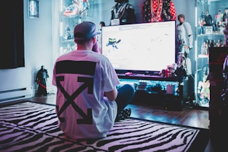 A person wearing a white t-shirt with a large black design is sitting cross-legged on a zebra-striped rug, facing a large screen in a room filled with action figures and collectibles displayed on illuminated shelves.