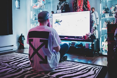A person wearing a white t-shirt with a large black design is sitting cross-legged on a zebra-striped rug, facing a large screen in a room filled with action figures and collectibles displayed on illuminated shelves.