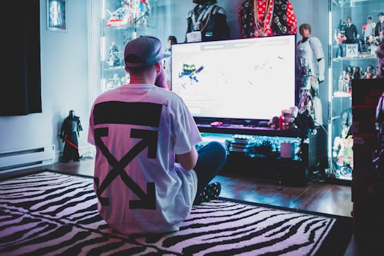 A person wearing a white t-shirt with a large black design is sitting cross-legged on a zebra-striped rug, facing a large screen in a room filled with action figures and collectibles displayed on illuminated shelves.