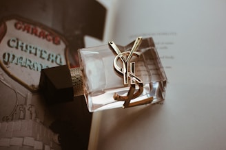 A close-up of a clear glass perfume bottle with gold accents and a black cap, placed on an open book. The background features a blurred sign with the words 'Chateau Marmont' visible, creating a vintage and luxurious ambiance.