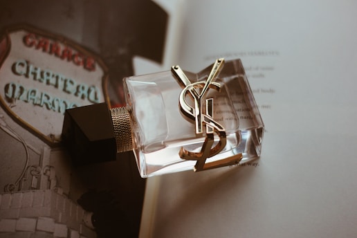 A close-up of a clear glass perfume bottle with gold accents and a black cap, placed on an open book. The background features a blurred sign with the words 'Chateau Marmont' visible, creating a vintage and luxurious ambiance.