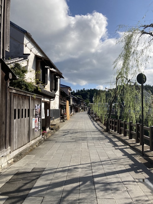 A quiet, traditional Japanese street is lined with old wooden buildings and willow trees. The sky is partly cloudy, allowing patches of sunlight to illuminate the pathway. Posters are visible on one of the buildings, adding a modern touch to the otherwise historic ambiance.