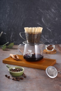 A pour-over coffee setup with a glass carafe labeled 'one two cups' filled with brewed coffee. On top of the carafe, there is a coffee dripper with a filter. The setting includes a wooden tray, a small green bowl with coffee beans, a wooden scoop with ground coffee, and a metal strainer. The background is a textured, dark surface.