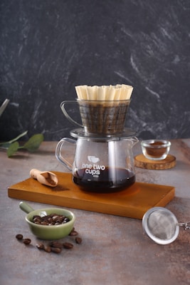 A pour-over coffee setup with a glass carafe labeled 'one two cups' filled with brewed coffee. On top of the carafe, there is a coffee dripper with a filter. The setting includes a wooden tray, a small green bowl with coffee beans, a wooden scoop with ground coffee, and a metal strainer. The background is a textured, dark surface.