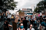 A large group of people is gathered on a city street for a protest. Many individuals are wearing masks and holding signs with messages advocating for justice and equality. The urban backdrop includes tall buildings and trees, under a cloudy sky.