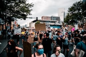A large group of people is gathered on a city street for a protest. Many individuals are wearing masks and holding signs with messages advocating for justice and equality. The urban backdrop includes tall buildings and trees, under a cloudy sky.