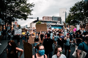 A large group of people is gathered on a city street for a protest. Many individuals are wearing masks and holding signs with messages advocating for justice and equality. The urban backdrop includes tall buildings and trees, under a cloudy sky.