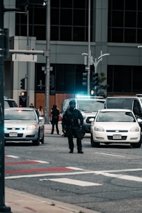 A heavily armed police officer stands in the middle of a street, surrounded by multiple vehicles including police cars with their lights on. The scene appears to be in an urban area with tall buildings in the background and several traffic signals. Pedestrians can be seen in the distance, indicating a possible law enforcement operation or checkpoint.