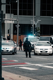 A heavily armed police officer stands in the middle of a street, surrounded by multiple vehicles including police cars with their lights on. The scene appears to be in an urban area with tall buildings in the background and several traffic signals. Pedestrians can be seen in the distance, indicating a possible law enforcement operation or checkpoint.