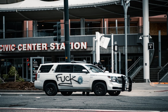 A white police vehicle is parked in front of a station labeled 'Civic Center Station.' The side of the vehicle is vandalized with graffiti. In the background, there are stairs, signage, and architectural details of the station entrance.