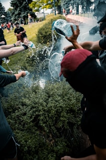 Several people are pouring water from bottles onto a smoky area surrounded by bushes in an outdoor setting. The group appears to be engaged in an activity that involves extinguishing smoke. There are other people and greenery visible in the background.