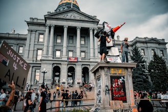 People are gathered in front of a large, historic building with columns and a dome. Many of them hold signs with various messages, and some are standing on a statue pedestal. The atmosphere suggests a protest or demonstration, with a focus on social justice themes.