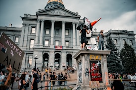People are gathered in front of a large, historic building with columns and a dome. Many of them hold signs with various messages, and some are standing on a statue pedestal. The atmosphere suggests a protest or demonstration, with a focus on social justice themes.