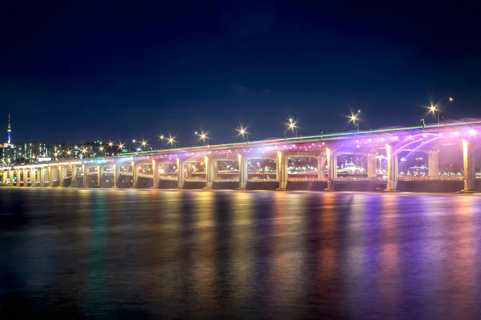Long illuminated bridge spans across water at night with colorful lights on structure