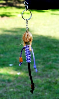 A handmade keychain with a knotted figure is suspended against a blurred green background. The figure has a face drawn on a wooden bead, and its body is made from purple and black cords.