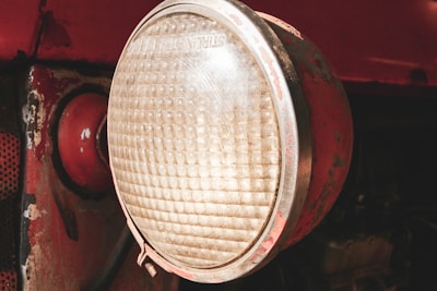 A close-up view of an old, weathered headlight attached to a vehicle. The headlight has a large, circular lens with a textured pattern, surrounded by a metal frame with visible rust and paint chipping. The surrounding area of the vehicle is painted red, with signs of wear and age.
