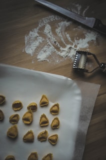 A wooden surface with scattered flour and a knife. A pasta cutter is nearby. Several pieces of handmade tortellini are arranged on parchment paper.