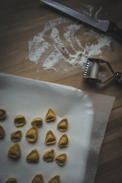 A wooden surface with scattered flour and a knife. A pasta cutter is nearby. Several pieces of handmade tortellini are arranged on parchment paper.