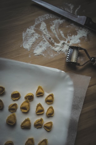 A wooden surface with scattered flour and a knife. A pasta cutter is nearby. Several pieces of handmade tortellini are arranged on parchment paper.