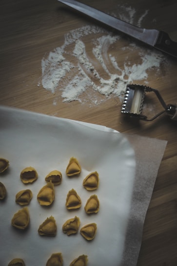 A wooden surface with scattered flour and a knife. A pasta cutter is nearby. Several pieces of handmade tortellini are arranged on parchment paper.