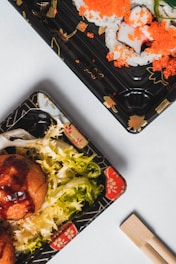 Two black plastic trays hold Japanese food items. One tray contains sushi rolls topped with bright orange fish roe, and the other tray features green leafy lettuce and what appears to be fried balls, likely takoyaki. Bamboo chopsticks are placed nearby on a white background.