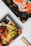 Two black plastic trays hold Japanese food items. One tray contains sushi rolls topped with bright orange fish roe, and the other tray features green leafy lettuce and what appears to be fried balls, likely takoyaki. Bamboo chopsticks are placed nearby on a white background.