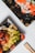 Two black plastic trays hold Japanese food items. One tray contains sushi rolls topped with bright orange fish roe, and the other tray features green leafy lettuce and what appears to be fried balls, likely takoyaki. Bamboo chopsticks are placed nearby on a white background.