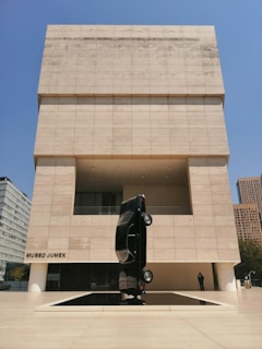 A modern art museum features a unique outdoor sculpture of a black car positioned vertically on its rear end. The museum's façade is composed of light-colored rectangular stone panels, contrasting with the bright blue sky. The building has a minimalist design with clean lines and geometric shapes.