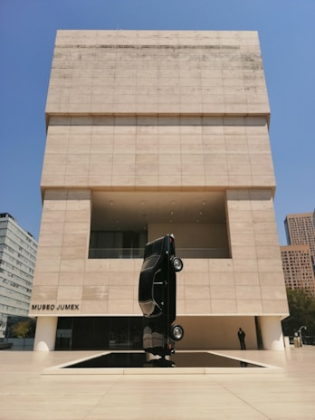 A modern art museum features a unique outdoor sculpture of a black car positioned vertically on its rear end. The museum's façade is composed of light-colored rectangular stone panels, contrasting with the bright blue sky. The building has a minimalist design with clean lines and geometric shapes.