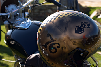 A motorcycle helmet with a golden-brown metallic finish features designs including a checkered pattern, dice, crossed wrenches, and the number 76. The helmet reflects a figure standing near a motorcycle, with grass and a fence in the background.