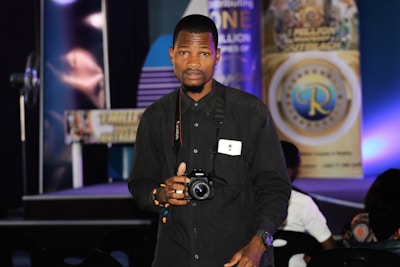 A person stands indoors holding a camera, looking directly at the viewer. They are wearing a black shirt and have a badge around their neck. Behind them, there are various banners with text and logos displayed on a stage, with blue and purple lighting adding color to the scene.